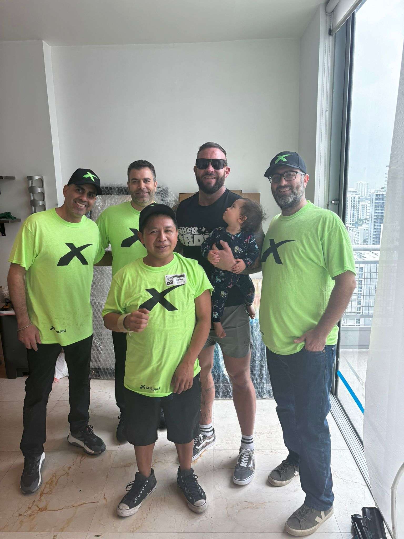 Four Xvanlines movers in neon green company shirts posing for a photo with a smiling male customer holding a young child in a modern apartment.
