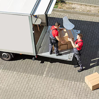 A mover in a green Xvanlines shirt stands on a green stepladder inside a moving truck, organizing "Lowe's" brand moving boxes and furniture wrapped in blue blankets and plastic.